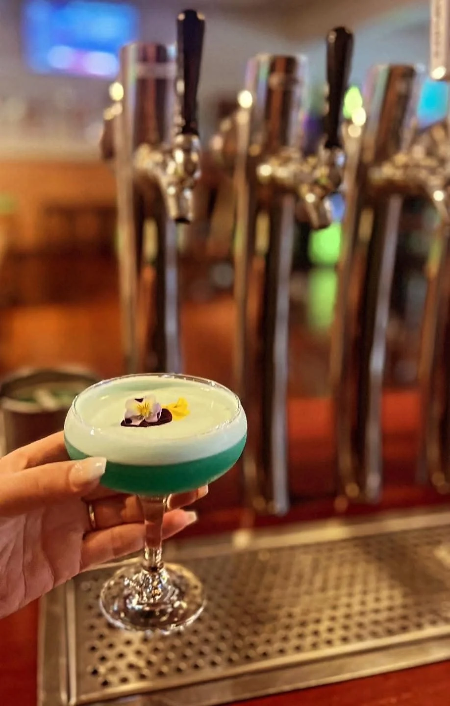 Teal craft cocktail with foam and edible flowers held in front of polished beer taps