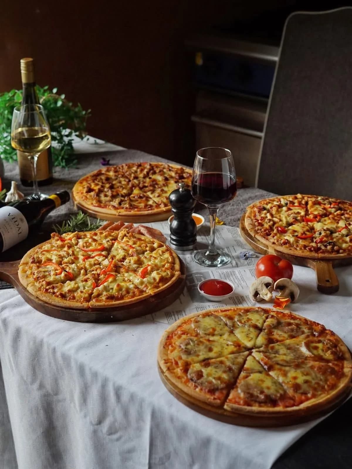 Four stone-baked pizzas on wooden paddles with wine bottles and fresh ingredients on a restaurant table